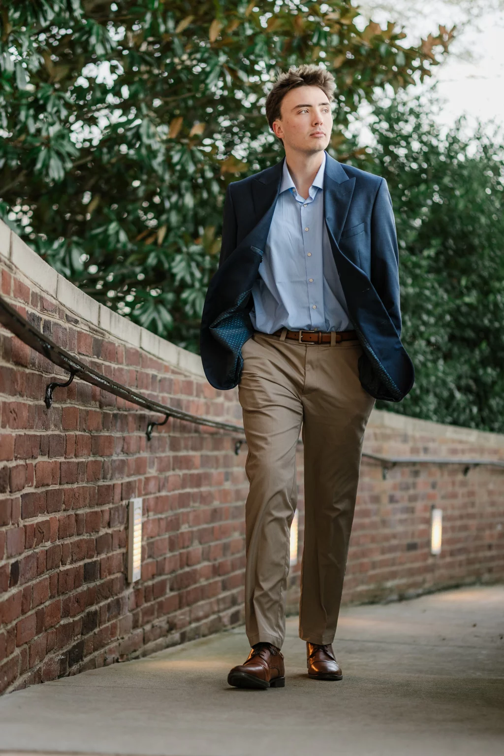 : A man in a navy blazer, light blue shirt, and khaki pants walks along a brick pathway with greenery in the background.