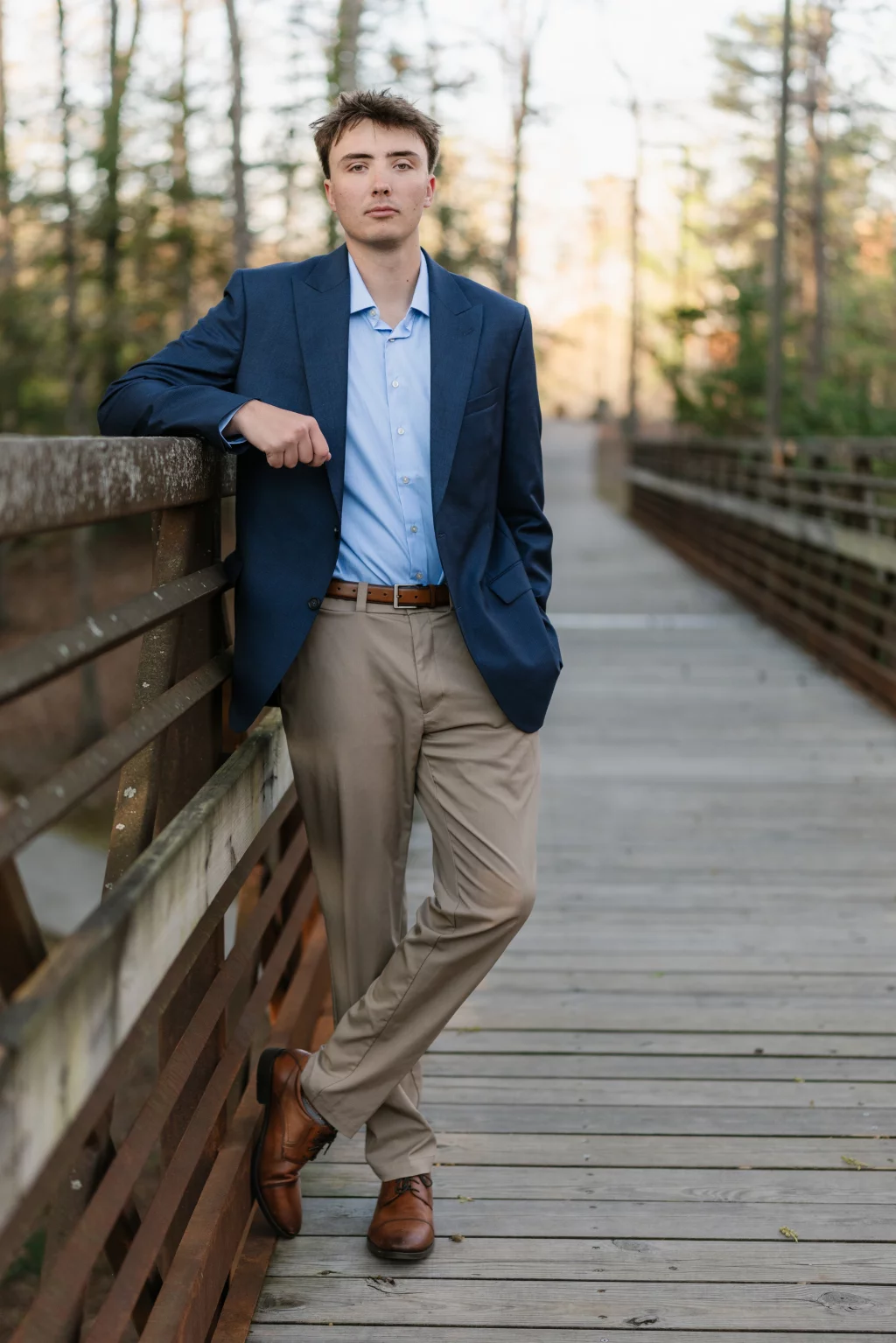 : Young man in a blue blazer, light blue shirt, and khaki pants stands on a wooden bridge in a wooded area, leaning against the railing with one hand in his pocket.
