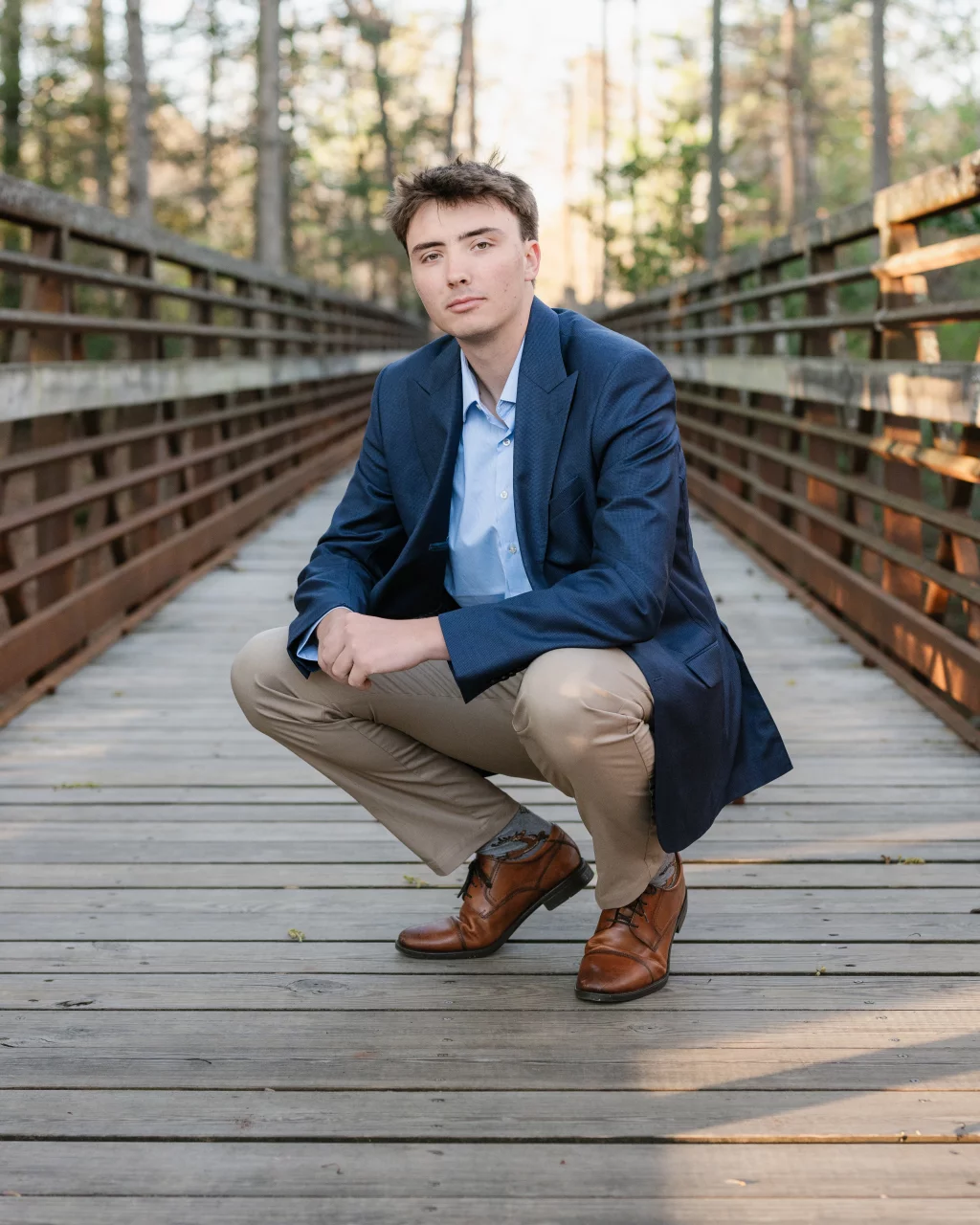 : A young man wearing a blue blazer, light blue shirt, khaki pants, and brown dress shoes squats on a wooden bridge in a forested area, looking at the camera.