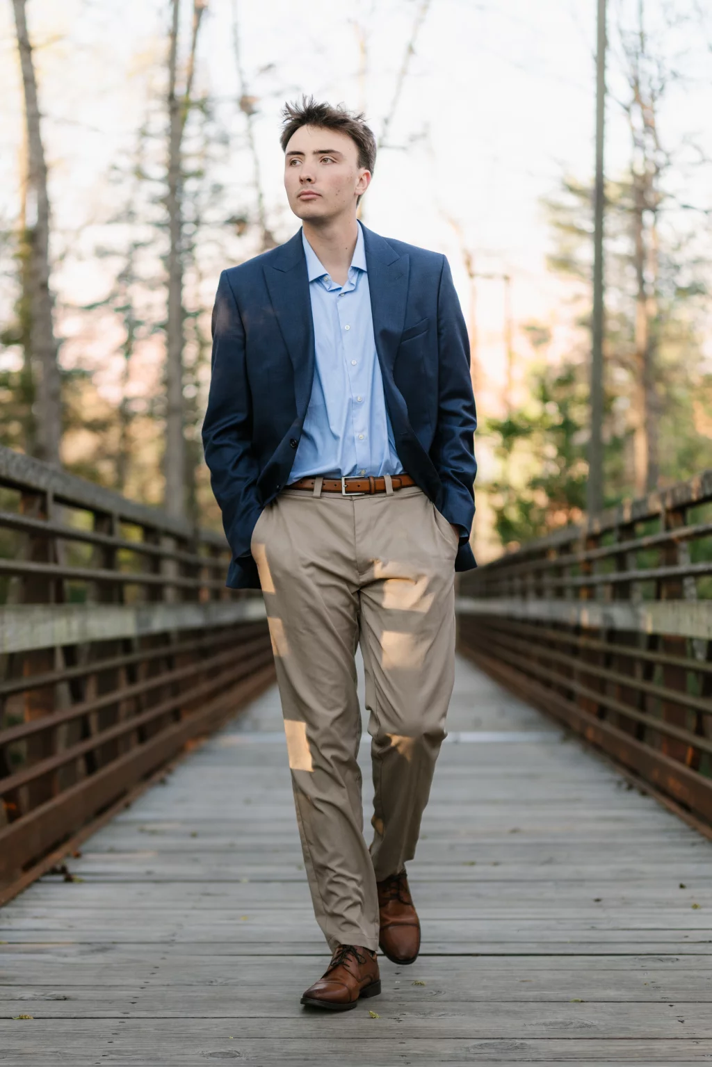 : A young man in a blue blazer, light blue shirt, khaki pants, and brown shoes walks on a wooden bridge with trees in the background.