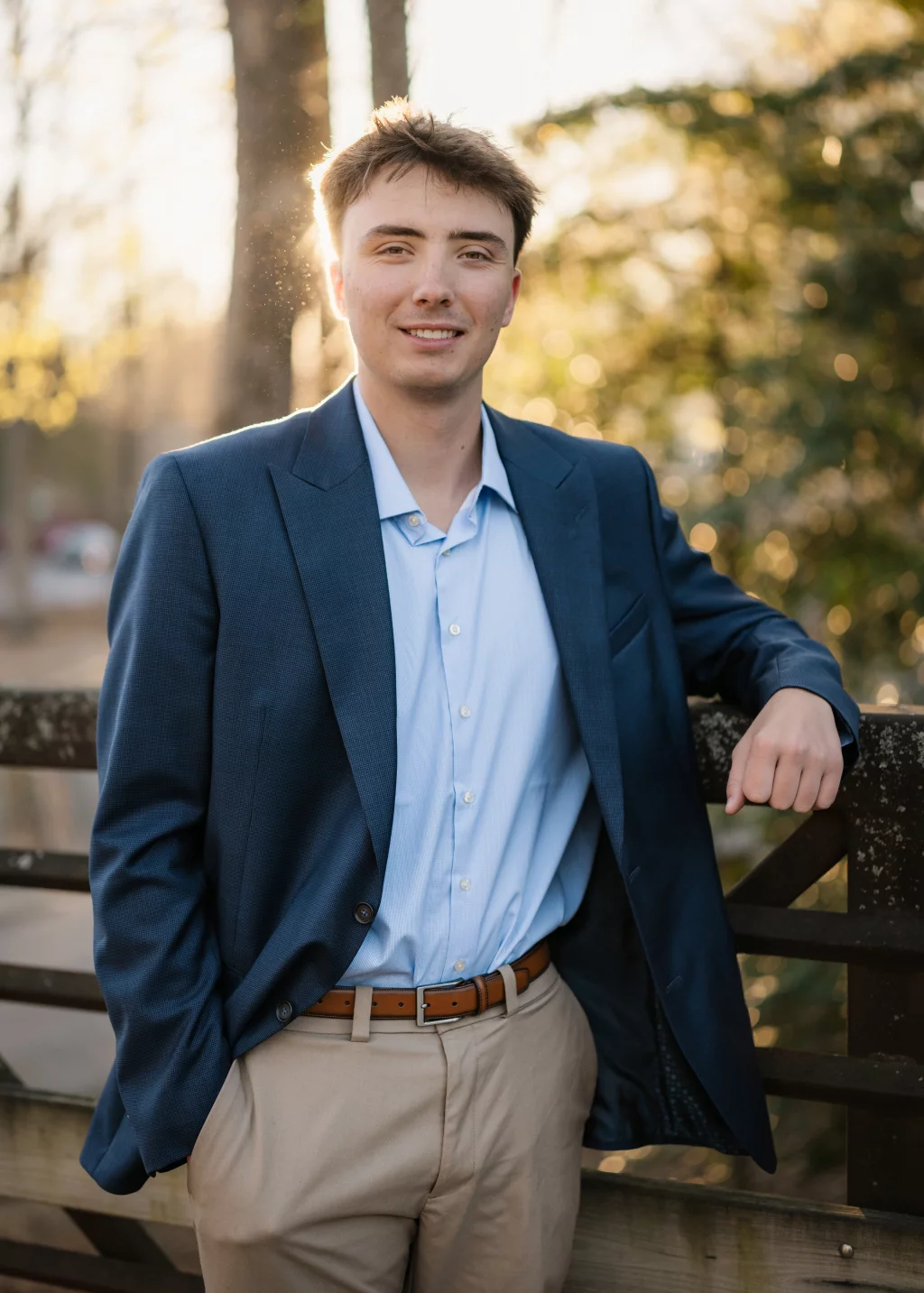 : Young man in a blue blazer and light blue shirt, standing outdoors by a wooden fence with trees and sunlight in the background, smiling at the camera.