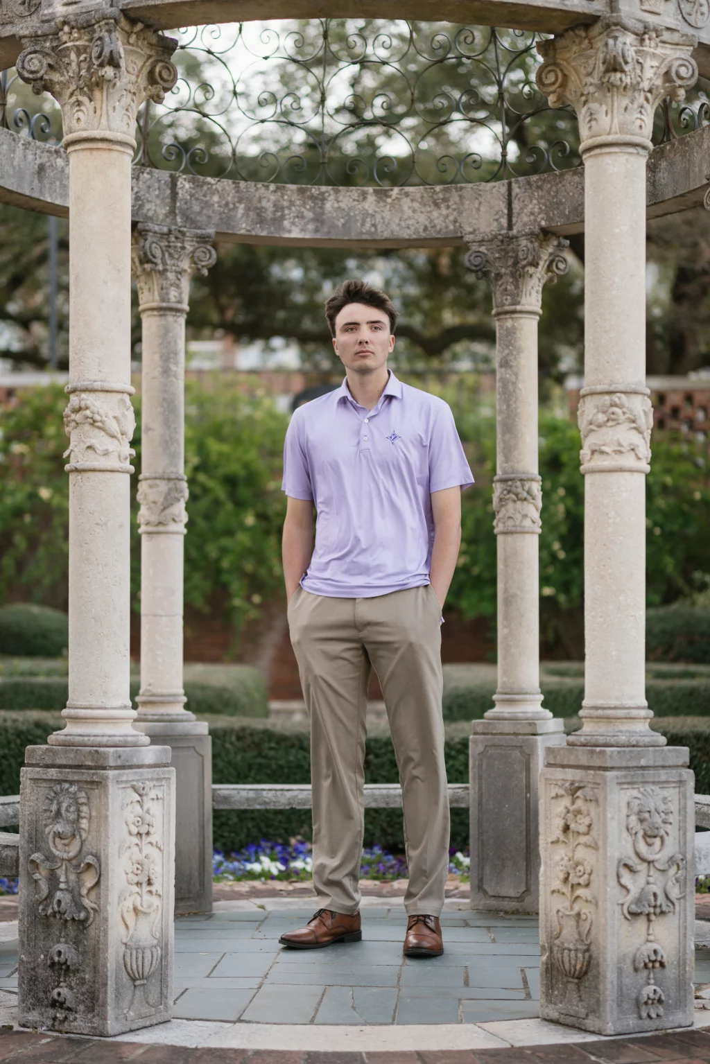 : A young man stands with hands in his pockets under a decorative stone gazebo with carved pillars, surrounded by greenery and flowers.