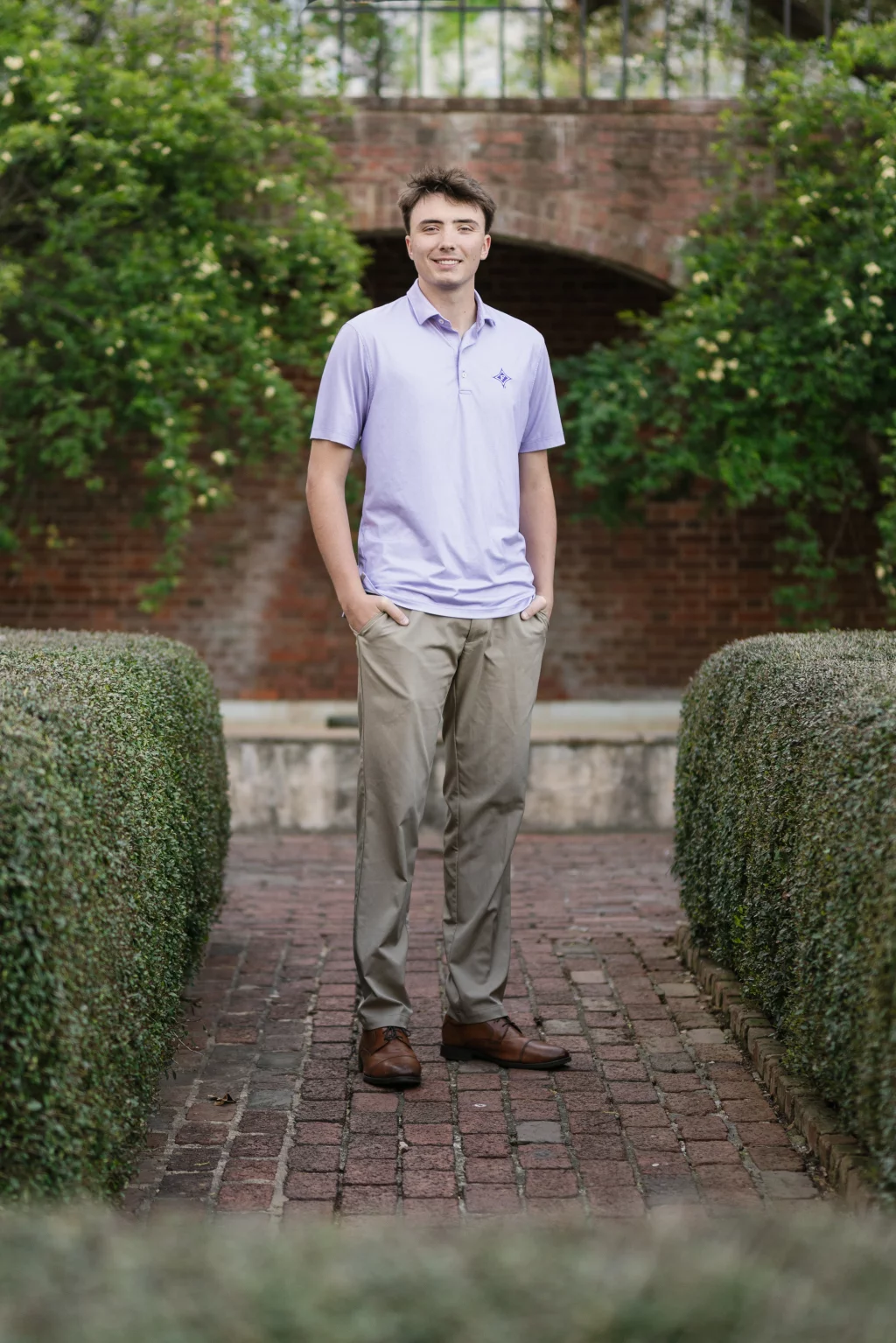 : Young man standing on a brick pathway lined with trimmed hedges, wearing a light purple polo shirt and khaki pants, with greenery and a brick archway in the background.