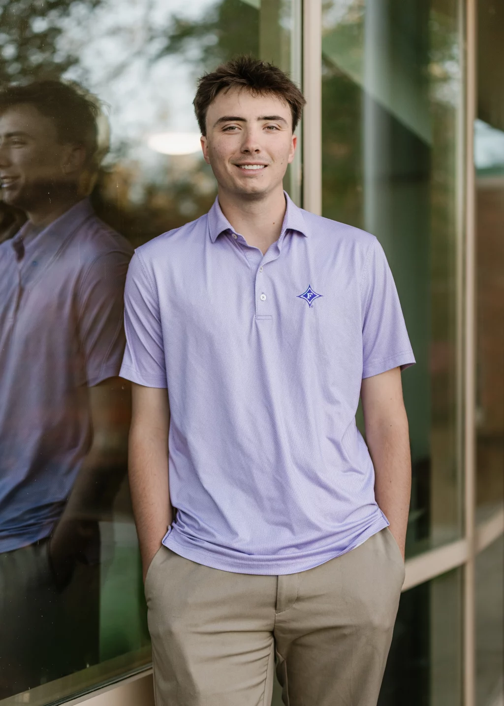 : A young man in a light purple polo shirt and khaki pants stands with hands in pockets, smiling and leaning against a glass wall with his reflection visible.