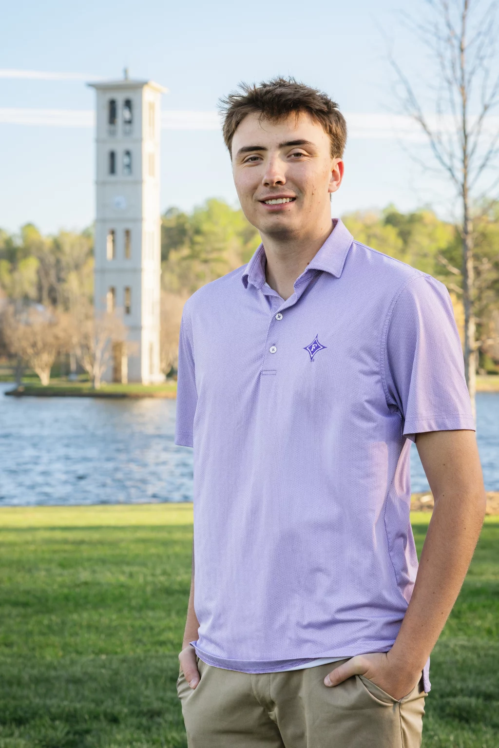 : Young man in a light purple polo shirt standing outdoors with a lake and a tall bell tower in the background.