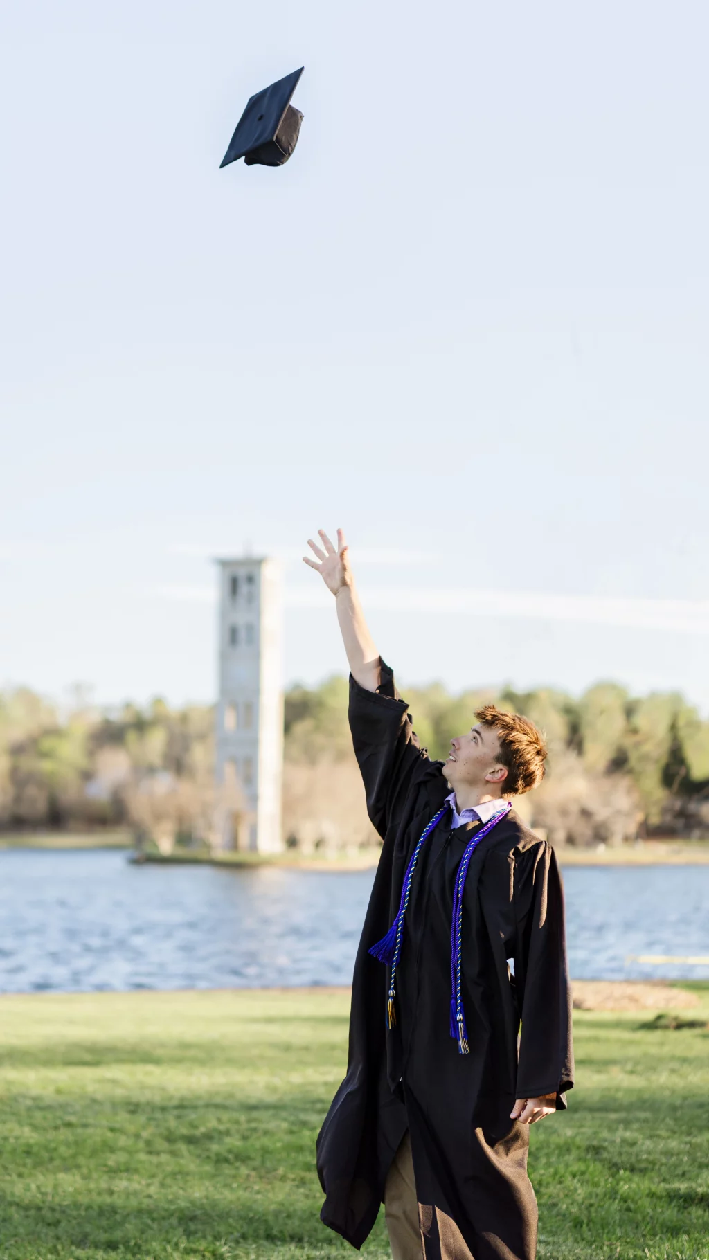 : A graduate in a black gown and honor cords stands on grass by a lake, reaching up as a graduation cap is thrown into the air. A tall clock tower is visible in the background.