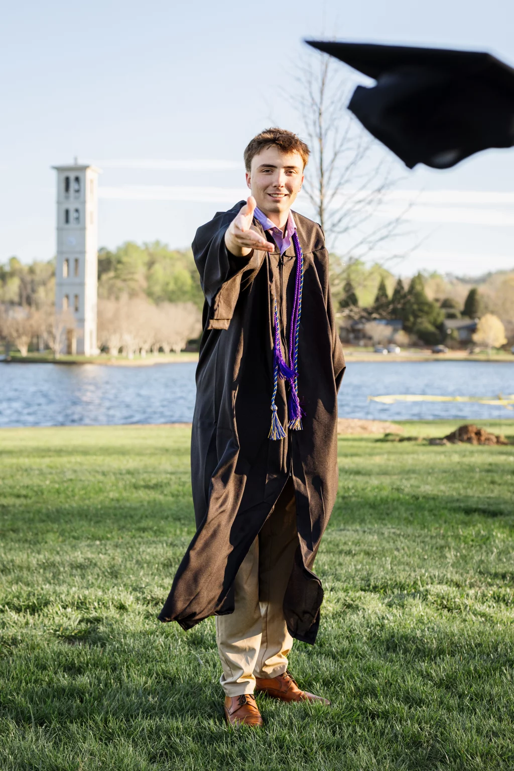 : A young man in a graduation gown stands on grass by a lake, tossing his graduation cap into the air, with a bell tower in the background.
