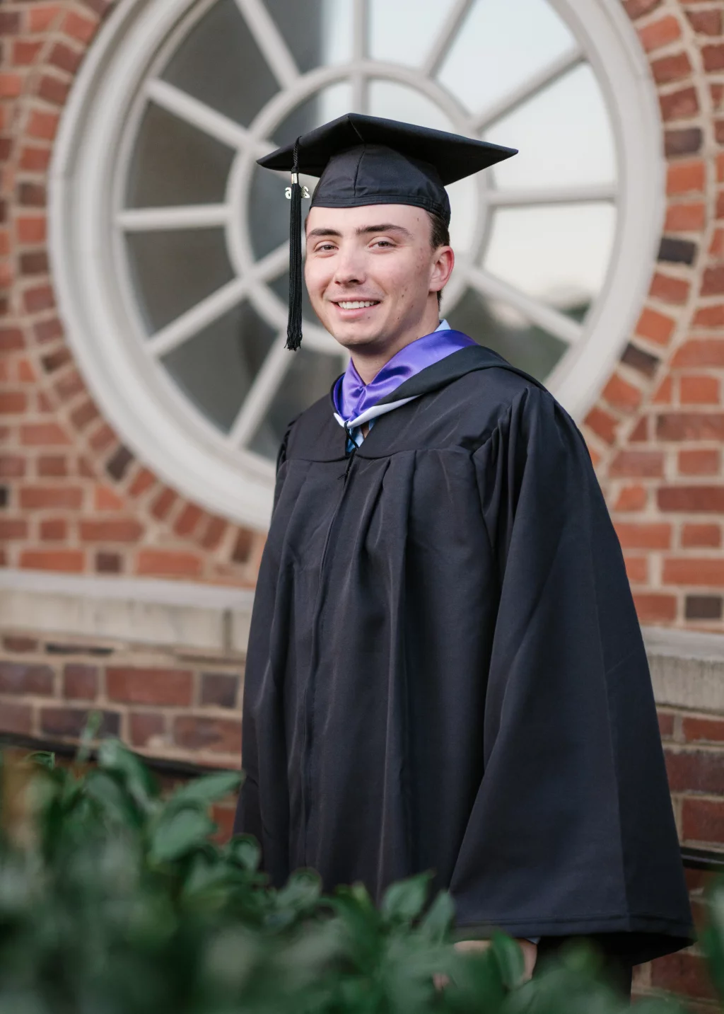 : A young man wearing a black graduation cap and gown stands in front of a brick building with a large circular window, smiling at the camera.