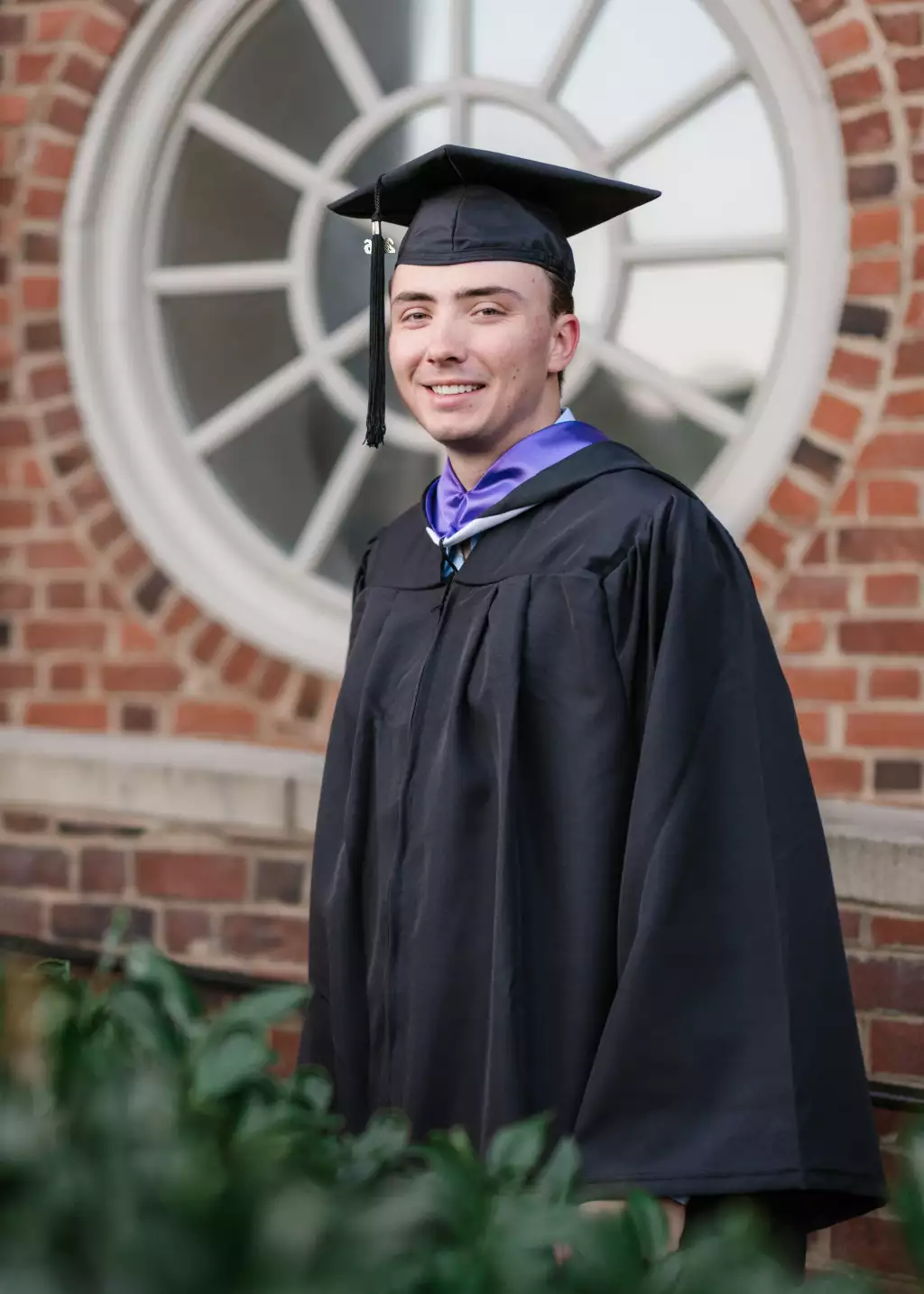 : A young man wearing a black graduation cap and gown stands in front of a brick building with a large circular window, smiling at the camera.