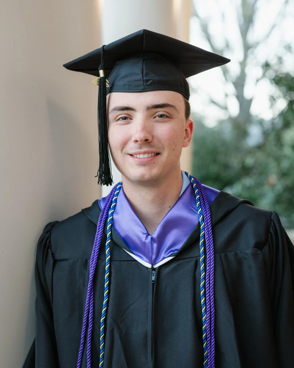 : Young man in a black graduation cap and gown with purple honor cords, standing and smiling near a column.