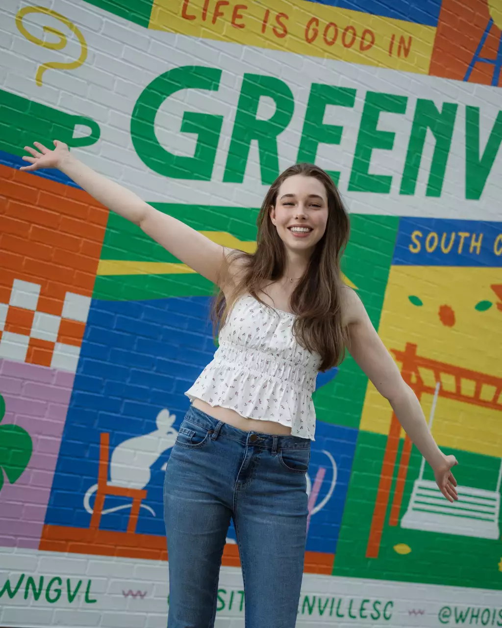 : Young woman with long brown hair smiling and spreading her arms wide in front of a colorful mural that reads "Life is good in Greenville". She is wearing a white sleeveless top with a small floral pattern and blue jeans.