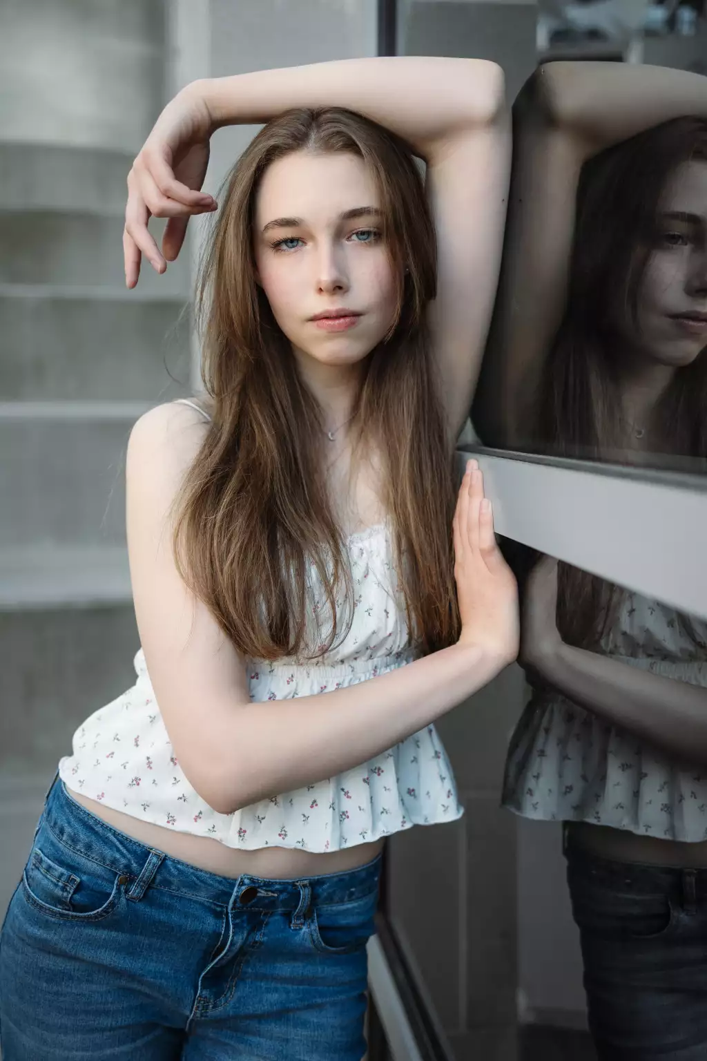 : Young woman with long brown hair, wearing a white floral sleeveless top and blue jeans, posing with one arm raised and resting on her head and the other hand touching a reflective glass surface, with her reflection visible, standing in front of a gray concrete background.
