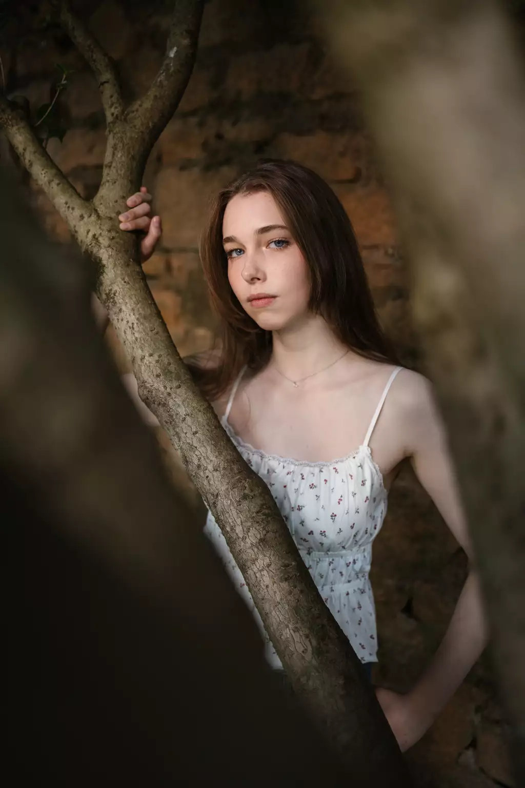 : Young woman with long brown hair and blue eyes standing behind tree branches, wearing a white floral sleeveless top, against a rustic stone wall background.