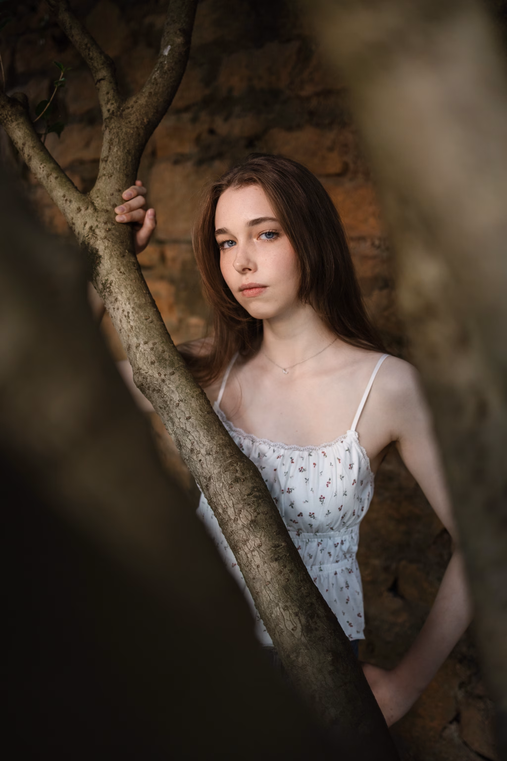 Abby's senior photos taken in downtown Greenville and Falls Park, Greenville, SC: Young woman with long brown hair and blue eyes standing behind tree branches, wearing a white floral sleeveless top, against a rustic stone wall background.