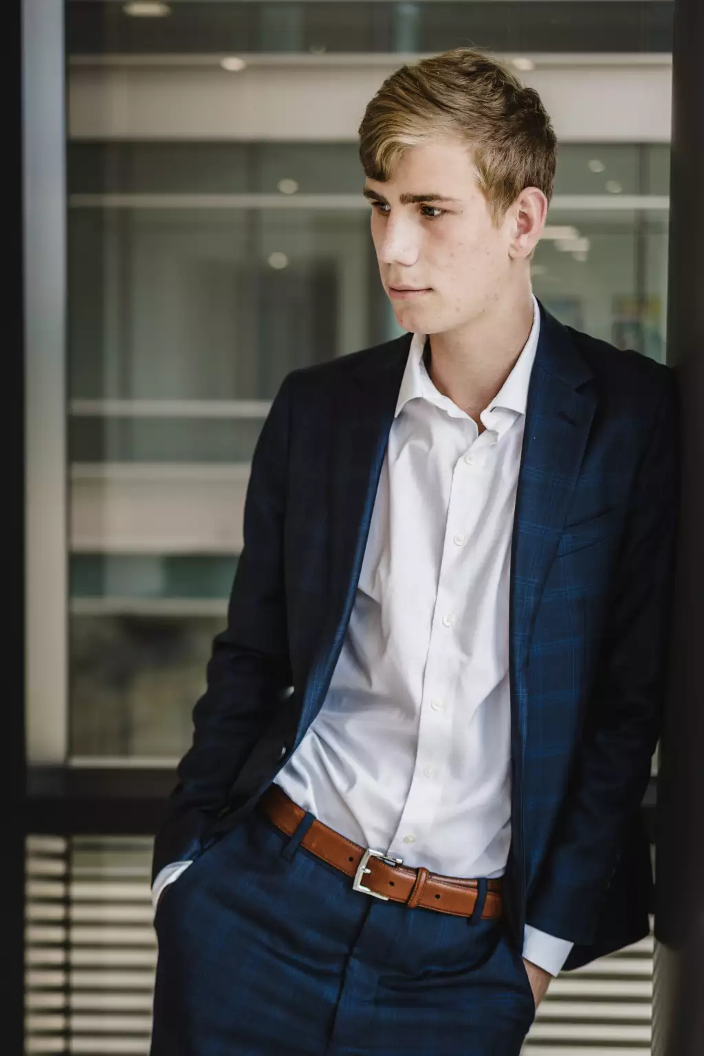 : Young man with light brown hair wearing a dark blue checkered suit and white shirt, standing indoors with hands in pockets, looking thoughtfully to the side.