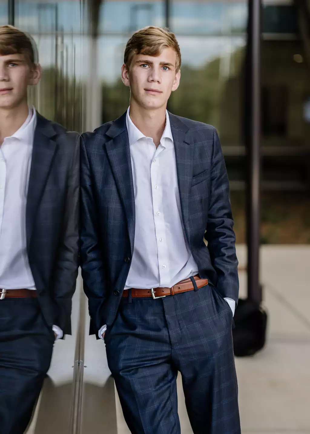 : Young man in a blue plaid suit with a white shirt and brown belt, leaning against a reflective glass wall with his hands in his pockets, looking directly at the camera.