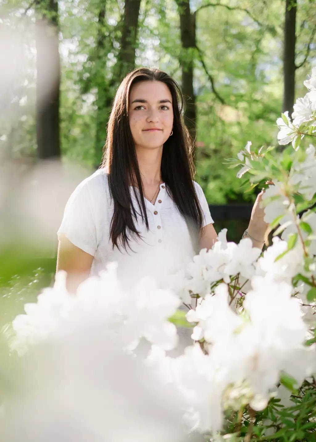 : Young woman with long dark hair wearing a white buttoned shirt standing outdoors among white flowering bushes with green trees in the background.