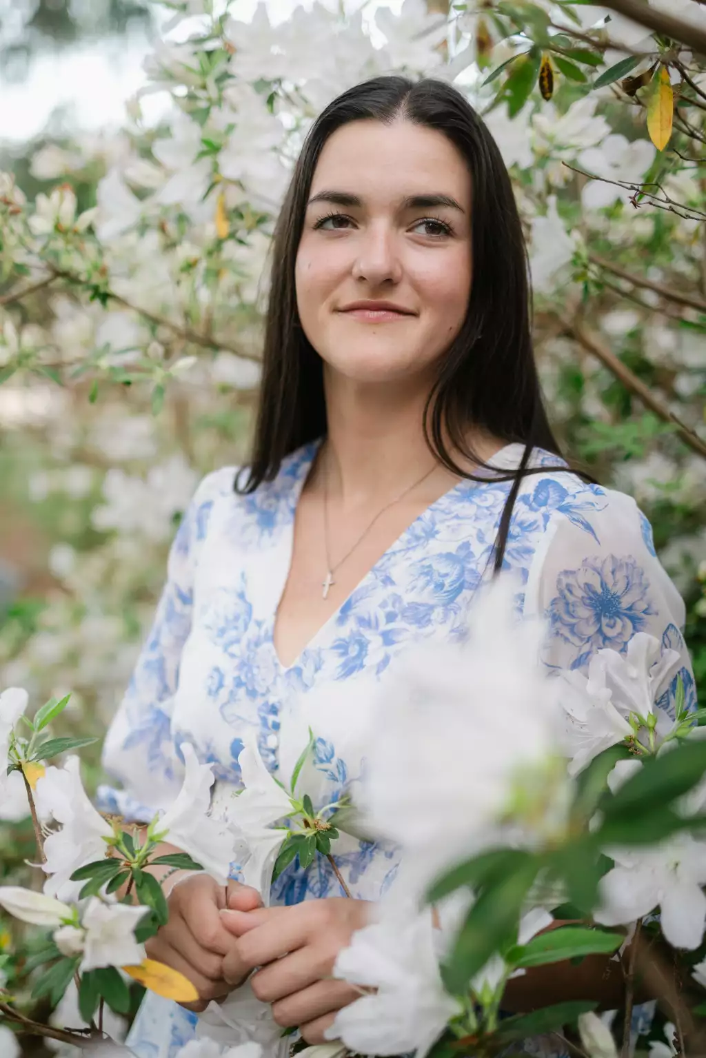 : Young woman with long dark hair wearing a white dress with blue floral patterns, standing among white flowering bushes, gently clasping her hands, with a neutral expression.