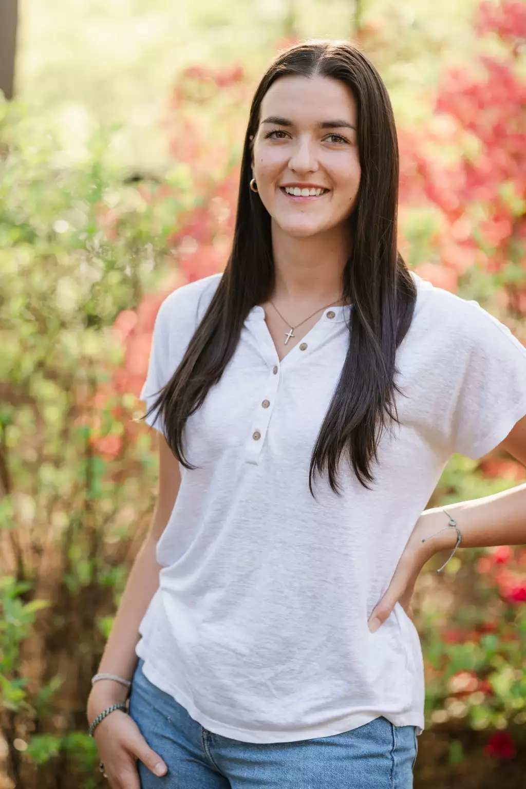 : Young woman with long dark hair smiling, wearing a white short-sleeve henley shirt, blue jeans, and a cross necklace, standing outdoors with a blurred background of green and red foliage.