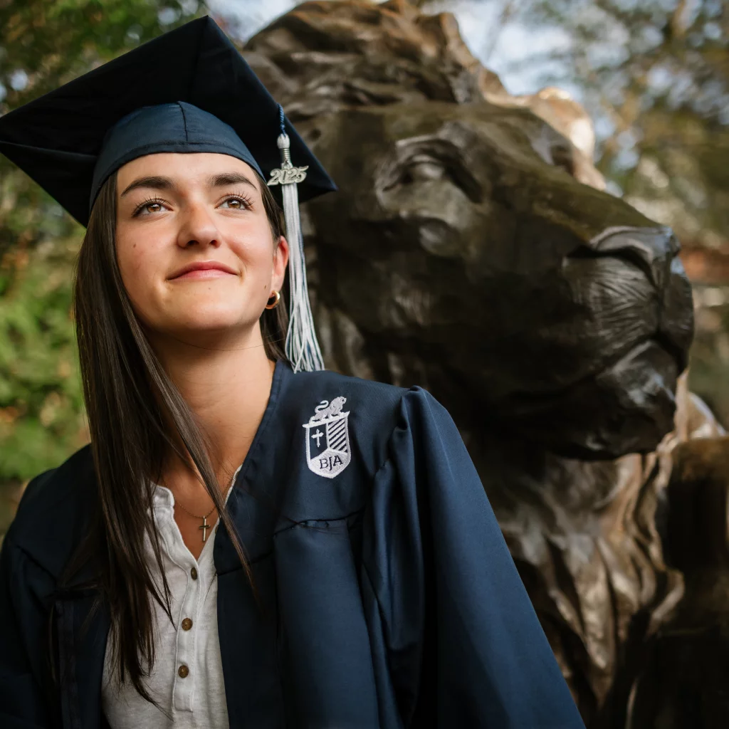 : Young woman in a navy blue graduation cap and gown with "2025" tassel standing next to a large bronze lion statue, looking upward with a slight smile.