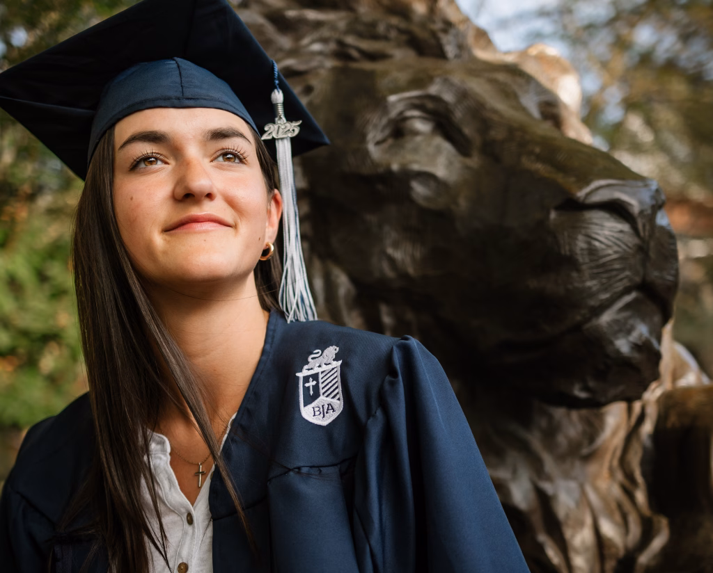 Annie's senior photos taken in downtown Greenville and Sue Simpson Garden, Greenville, SC: Young woman in a blue graduation gown and cap with a "2025" tassel, standing next to a large lion statue outdoors with a thoughtful expression.