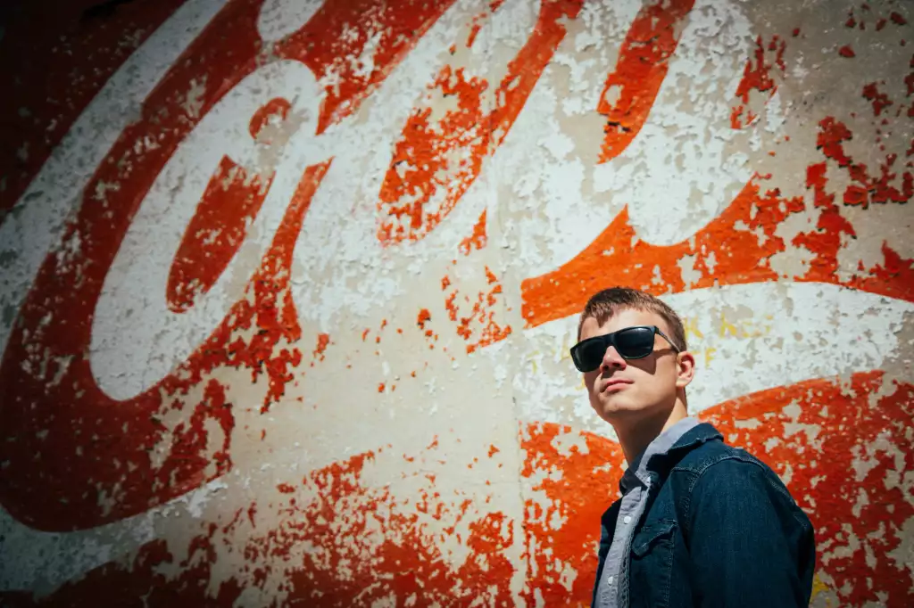 Cayden's senior photos taken downtown Greenville SC: Young man wearing dark sunglasses and a denim jacket standing in front of a large, weathered red and white Coca-Cola mural.