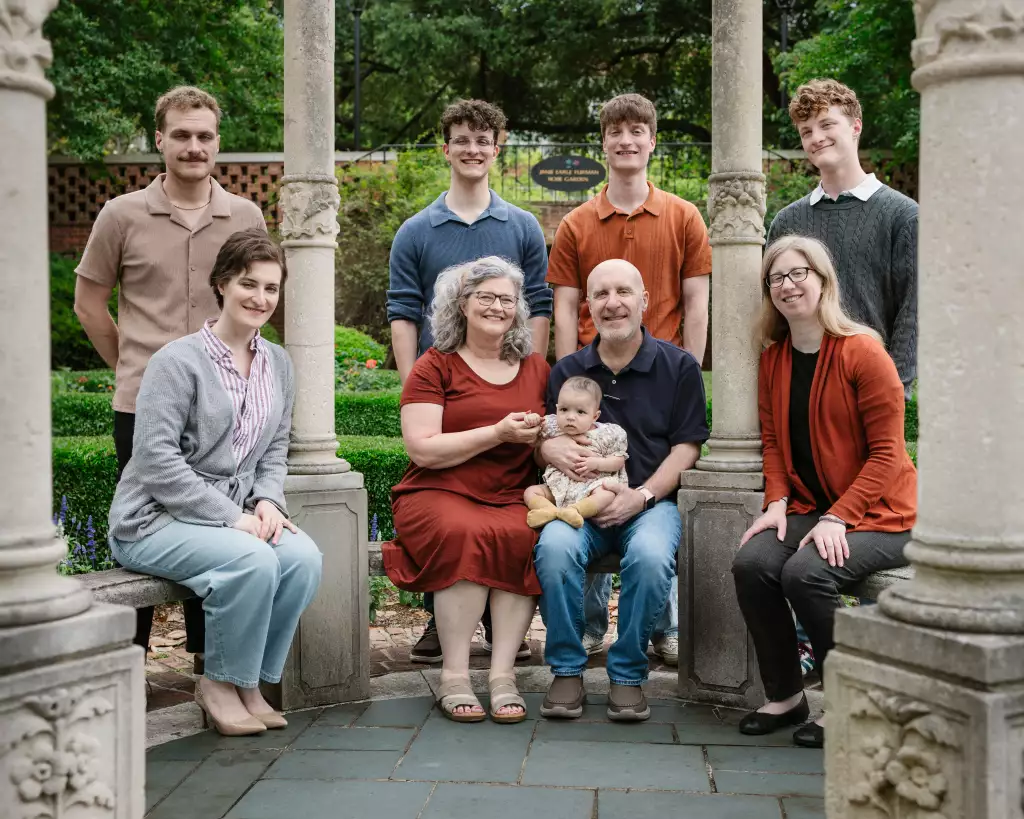 : Group portrait of nine people in a garden setting framed by stone pillars, featuring three older adults seated in the center, holding a baby, with three younger men standing behind them and three adults seated on stone benches on either side.