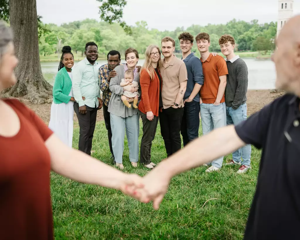 : A diverse group of ten people, including one holding a baby, standing outdoors on grass near a lake with trees in the background. In the foreground, two blurred adults hold hands, framing the group behind them.