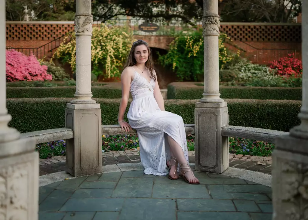 : Young woman in a white dress and sandals sitting on a stone bench between ornate stone columns in a garden with blooming flowers and trimmed hedges.