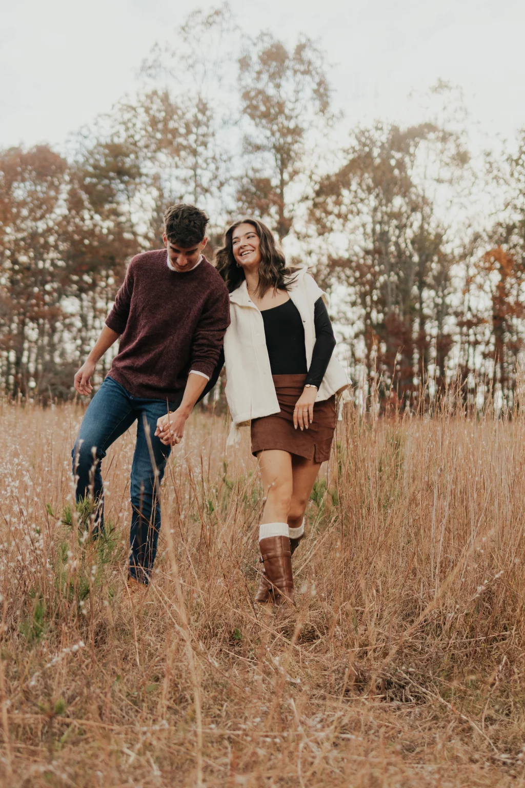 Ethan & Savanah's couple photo session taken at Campbell's Covered Bride, Greenville County: Young couple holding hands and walking through a tall dry grass field with autumn trees in the background.