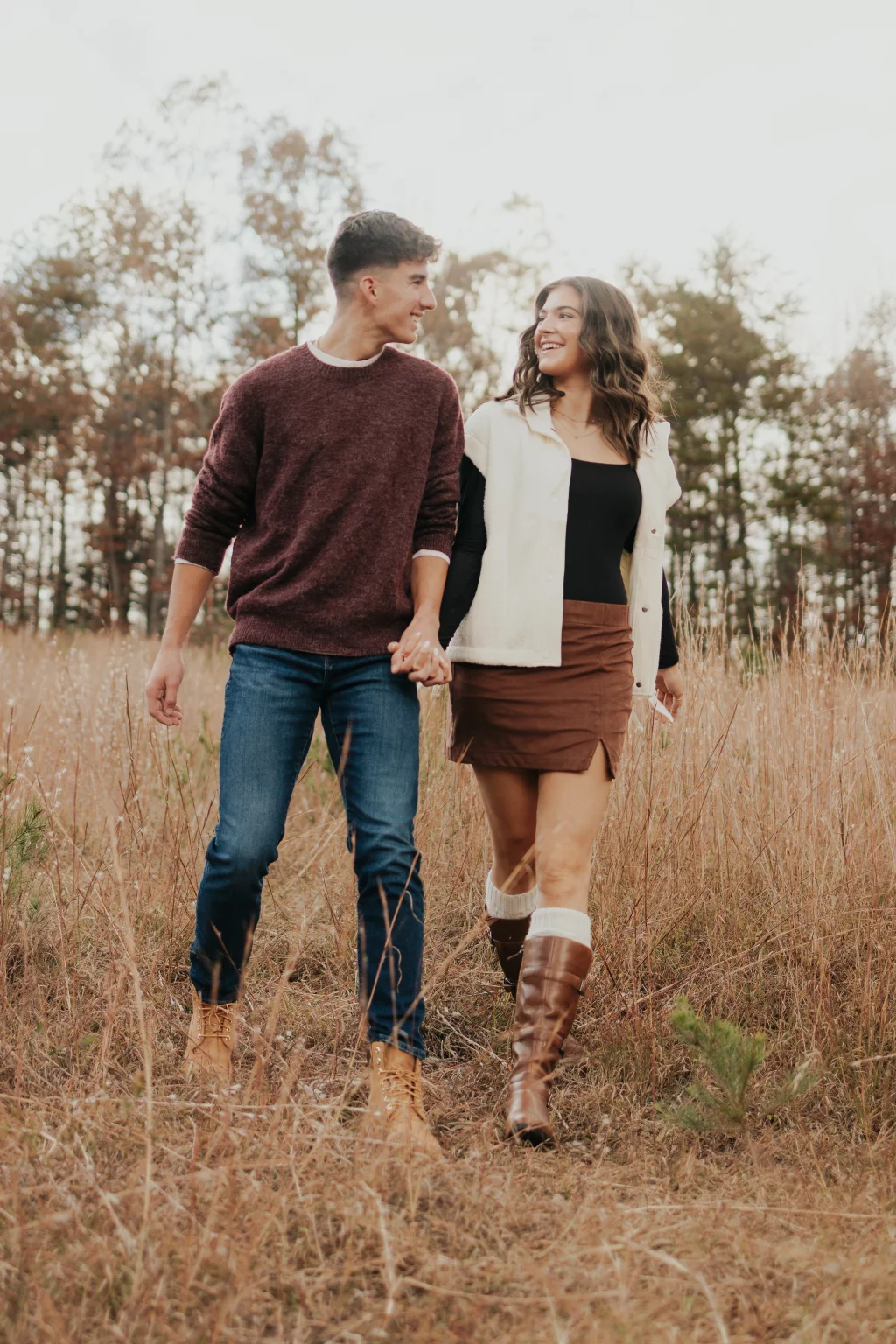 Ethan & Savanah's couple photo session taken at Campbell's Covered Bride, Greenville County: Couple holding hands walking through a dry grassy field with trees in the background, smiling at each other.