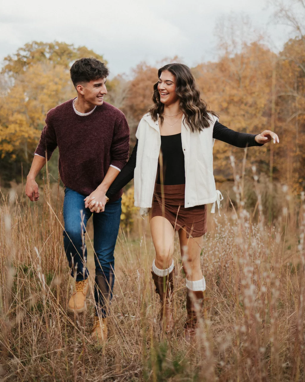 Ethan & Savanah's couple photo session taken at Campbell's Covered Bride, Greenville County: Young couple holding hands walking through tall grass in autumn, smiling at each other.