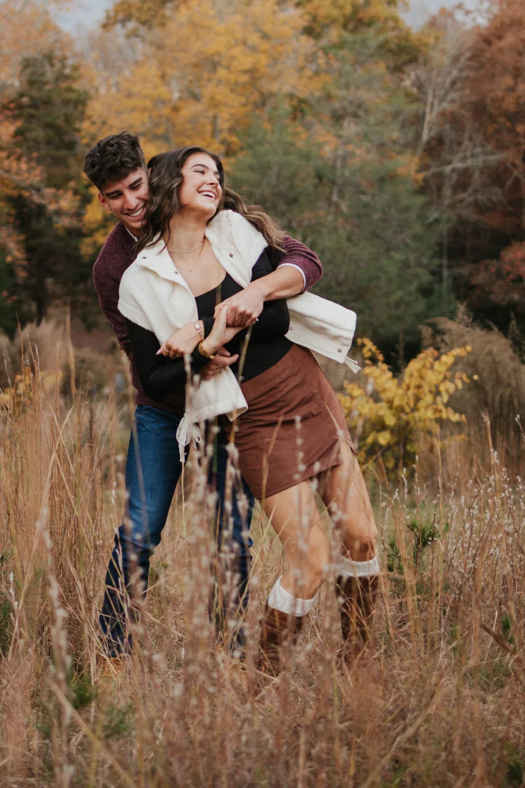 Ethan & Savanah's couple photo session taken at Campbell's Covered Bride, Greenville County: Young couple laughing and embracing in a tall grassy field with autumn trees in the background.