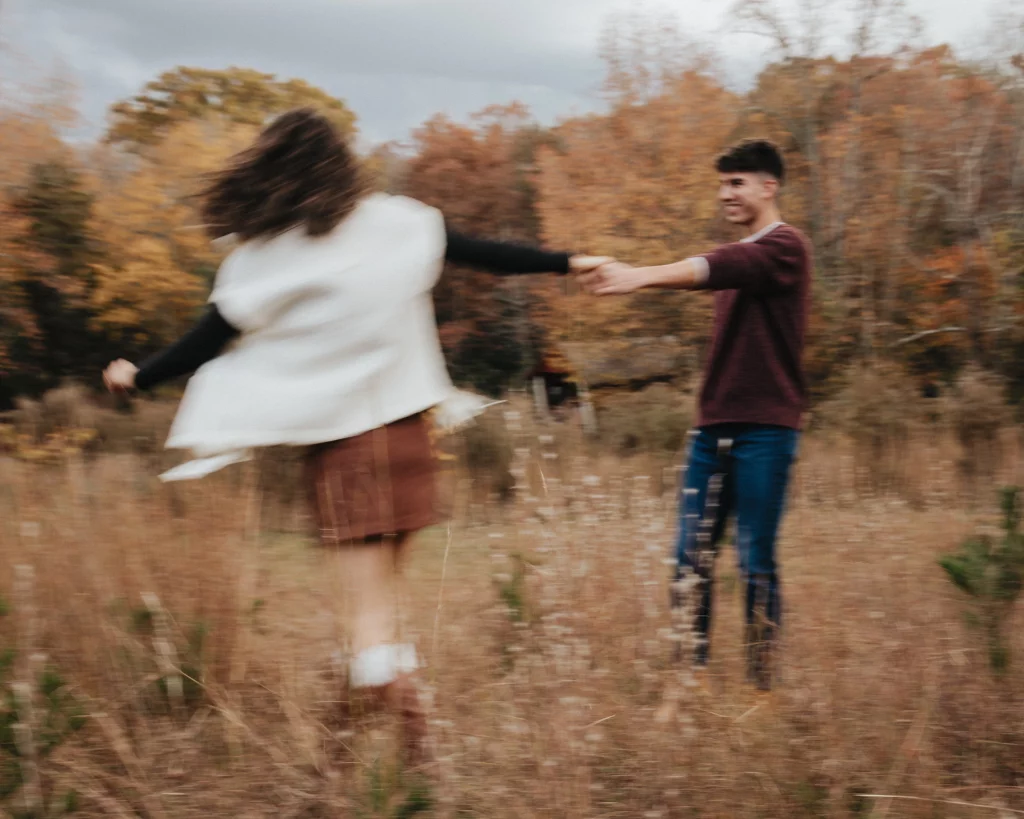 Ethan & Savanah's couple photo session taken at Campbell's Covered Bride, Greenville County: Two people holding hands and spinning in a grassy field with autumn trees in the background, image slightly blurred.