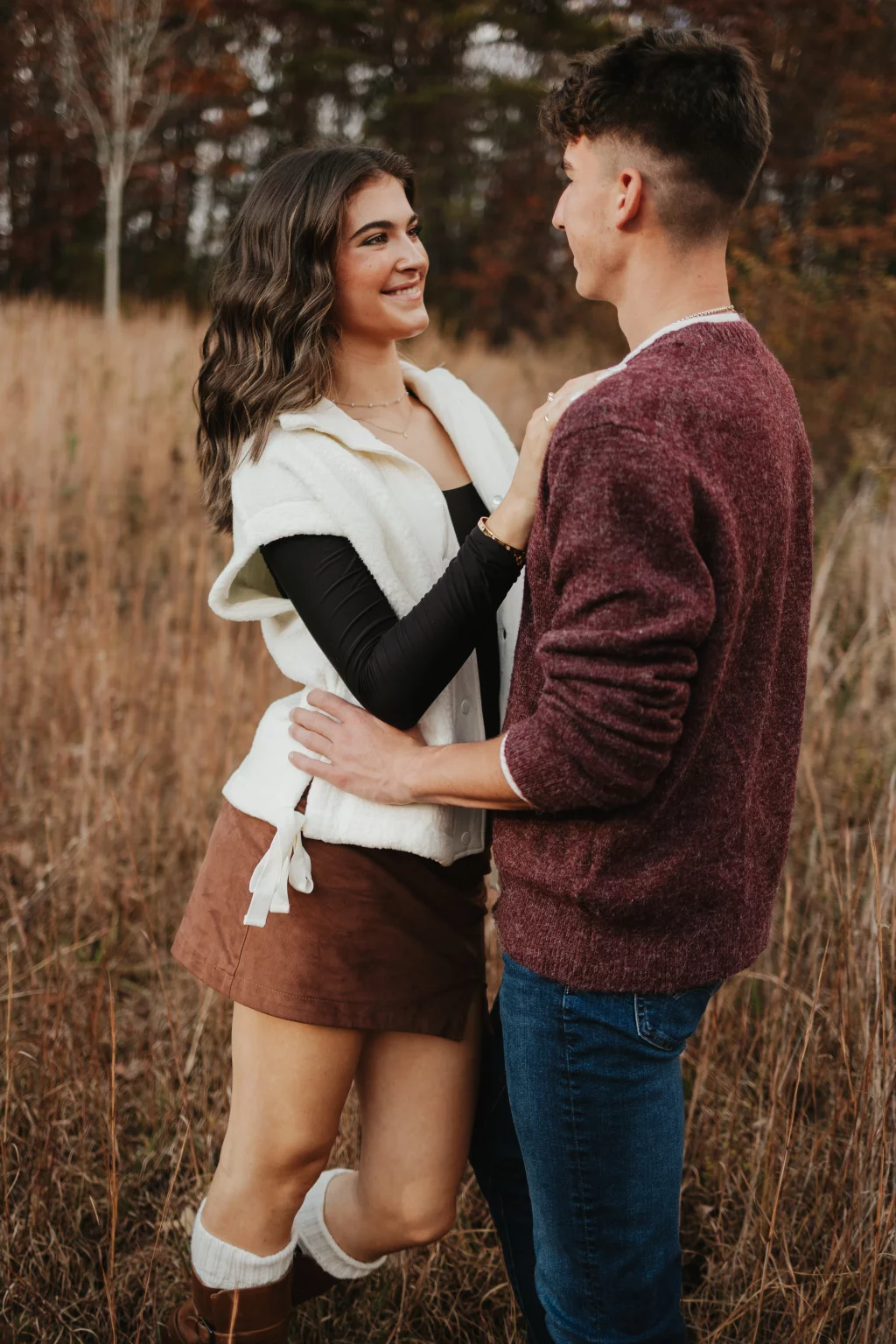 Ethan & Savanah's couple photo session taken at Campbell's Covered Bride, Greenville County: Young couple smiling at each other outdoors in a field with autumn foliage, the woman wearing a brown skirt and white vest, and the man in a maroon sweater and jeans.