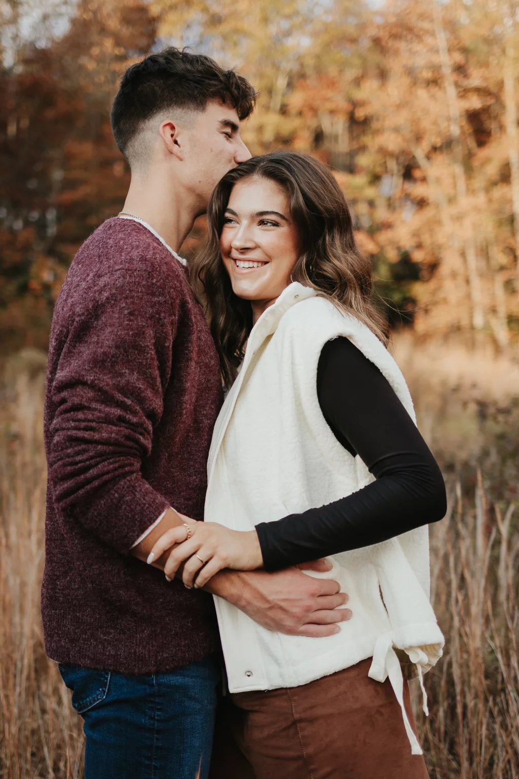 Ethan & Savanah's couple photo session taken at Campbell's Covered Bride, Greenville County: Young couple embracing outdoors in autumn, man in maroon sweater kissing woman's forehead, woman smiling in white vest.