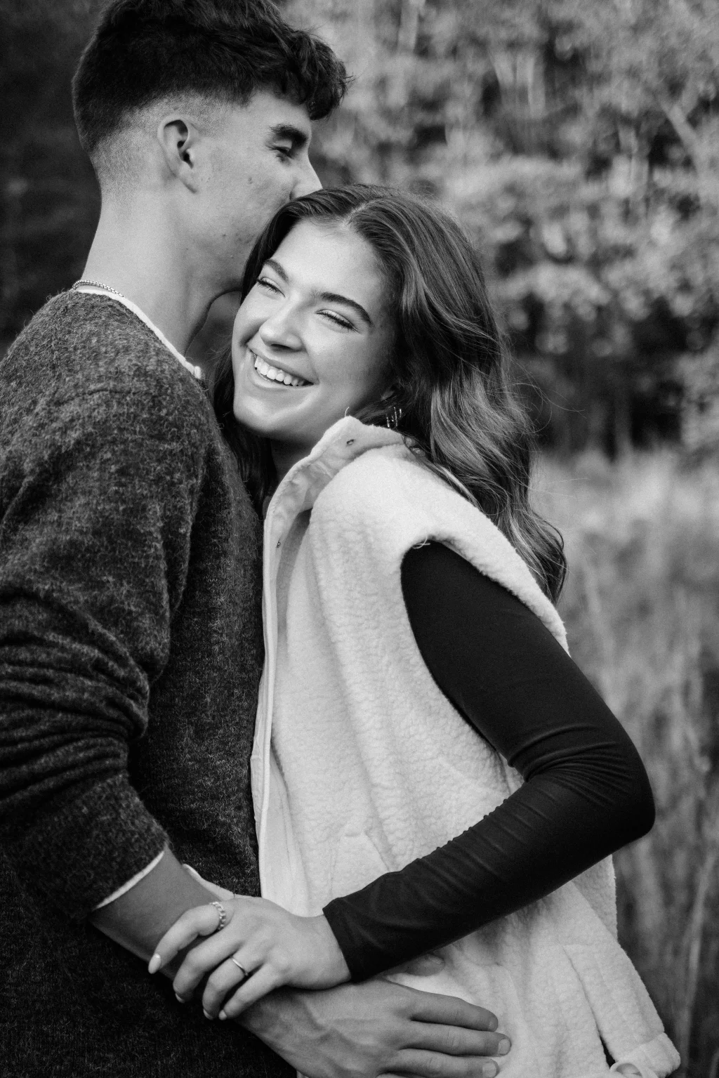 Ethan & Savanah's couple photo session taken at Campbell's Covered Bride, Greenville County: Black and white photo of a smiling woman hugging a man from the front as he leans in close, holding hands outdoors.