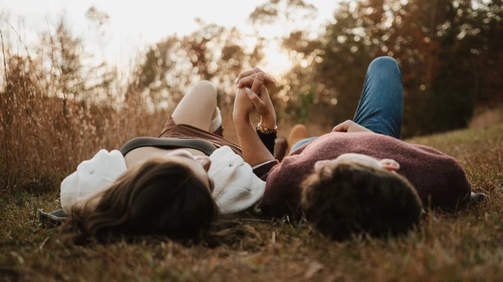 Ethan & Savanah's couple photo session taken at Campbell's Covered Bride, Greenville County: Two people lying on grass in a field holding hands with a sunset and trees in the background.