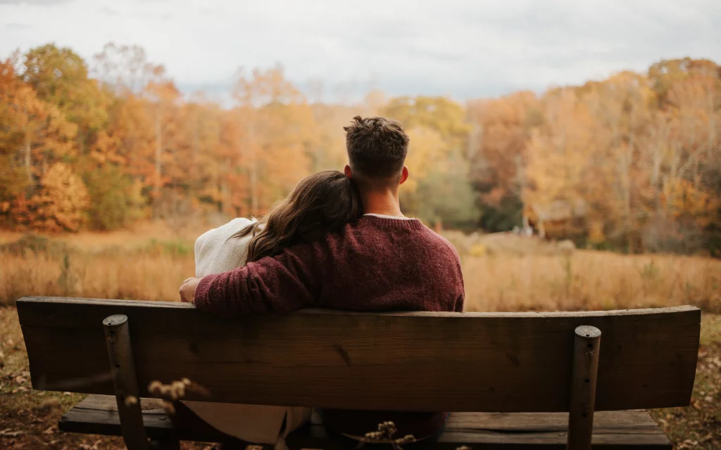 Ethan & Savanah's couple photo session taken at Campbell's Covered Bride, Greenville County: Couple sitting on a wooden bench in an autumn park, woman resting her head on man's shoulder, surrounded by fall foliage.