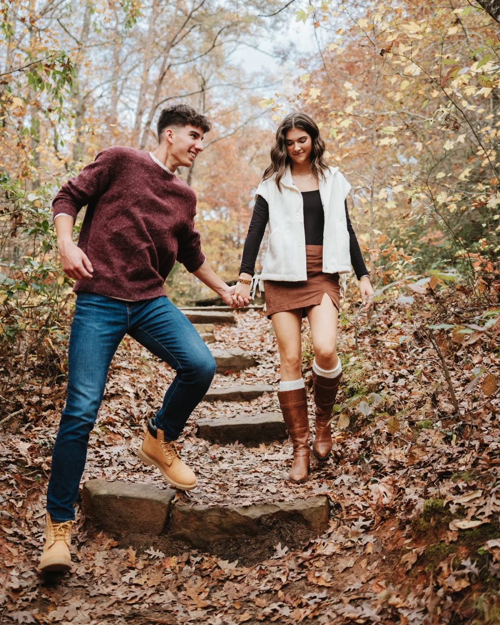 Ethan & Savanah's couple photo session taken at Campbell's Covered Bride, Greenville County: Young couple holding hands and walking down a leaf-covered stone path in a forest during autumn.