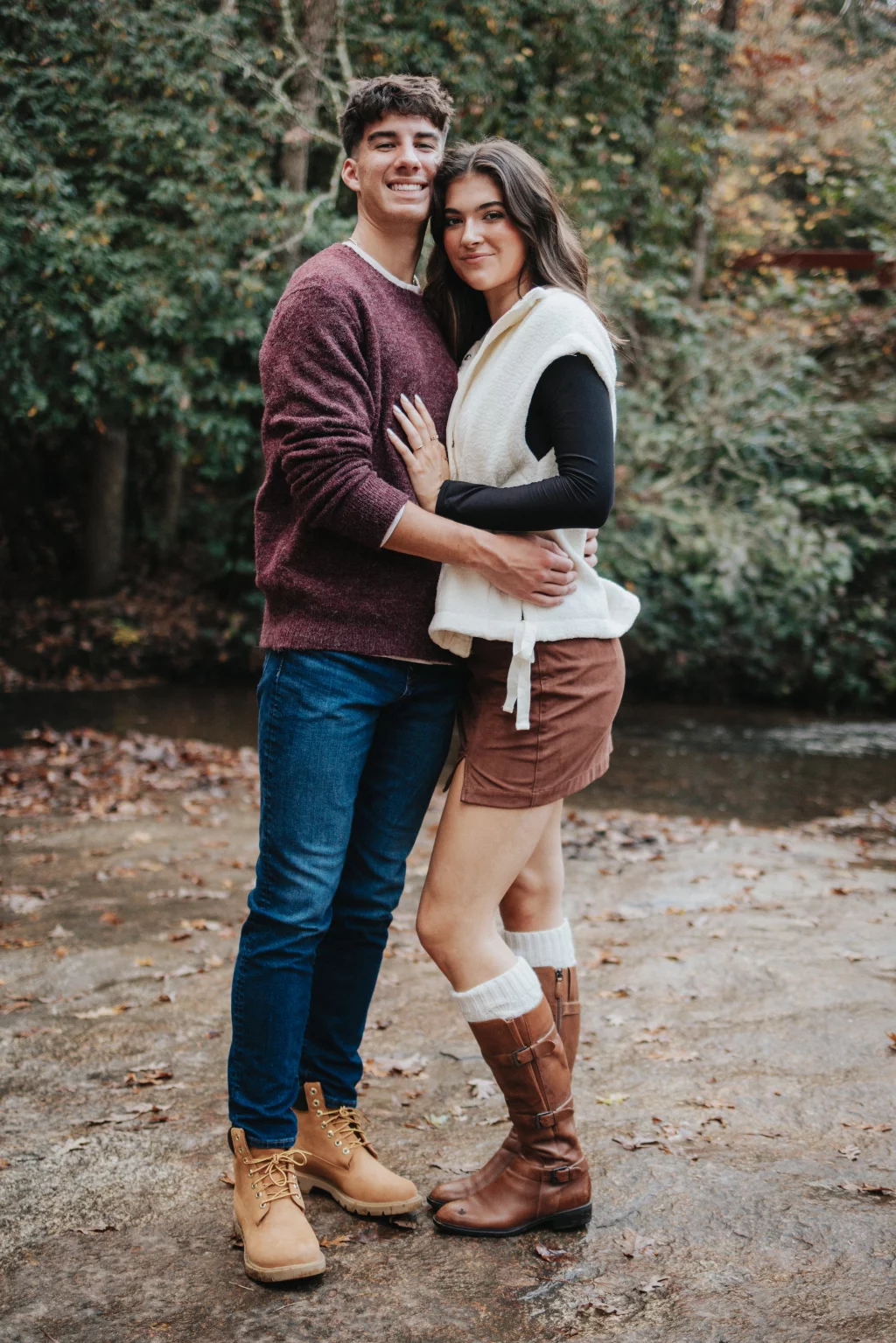 Ethan & Savanah's couple photo session taken at Campbell's Covered Bride, Greenville County: Young couple standing outdoors by a creek in autumn, embracing and smiling at the camera, surrounded by trees with fall foliage.
