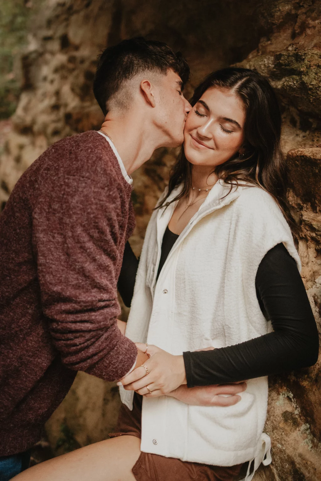 Ethan & Savanah's couple photo session taken at Campbell's Covered Bride, Greenville County: Young man kissing young woman’s cheek while holding hands, both smiling, against a rustic stone wall.