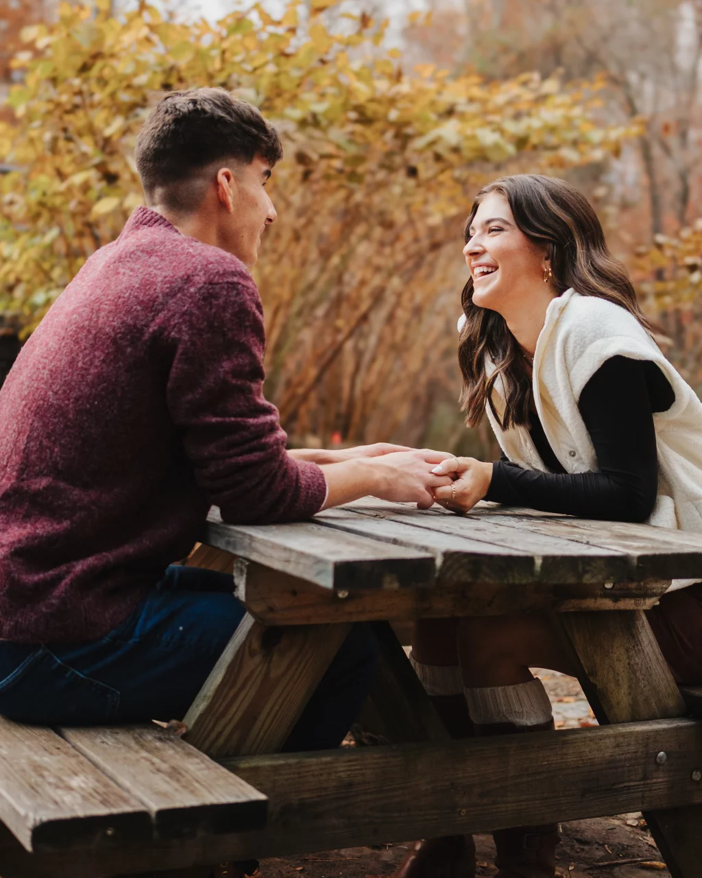 Ethan & Savanah's couple photo session taken at Campbell's Covered Bride, Greenville County: Young couple holding hands and laughing while sitting opposite each other at a wooden picnic table outdoors in autumn.