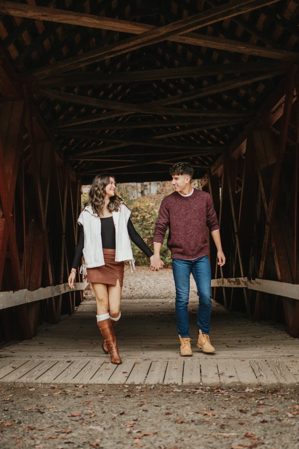 Ethan & Savanah's couple photo session taken at Campbell's Covered Bride, Greenville County: Couple holding hands and smiling at each other while walking through a wooden covered bridge in autumn.