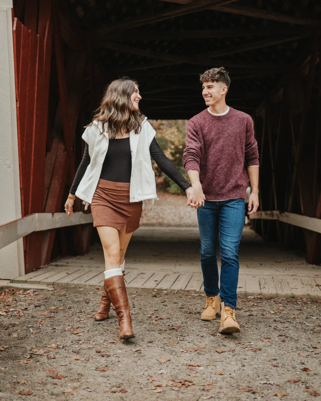 Ethan & Savanah's couple photo session taken at Campbell's Covered Bride, Greenville County: Young couple holding hands and walking together on a rustic covered bridge in autumn.