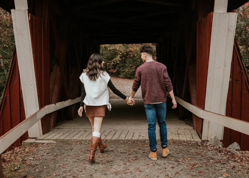 Ethan & Savanah's couple photo session taken at Campbell's Covered Bride, Greenville County: Couple holding hands walking into a covered wooden bridge surrounded by autumn foliage.