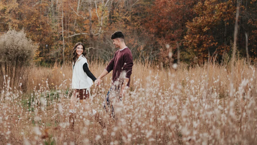 Ethan & Savanah's couple photo session taken at Campbell's Covered Bride, Greenville County: Young couple holding hands and walking through a field of tall grasses and wildflowers with autumn trees in the background.