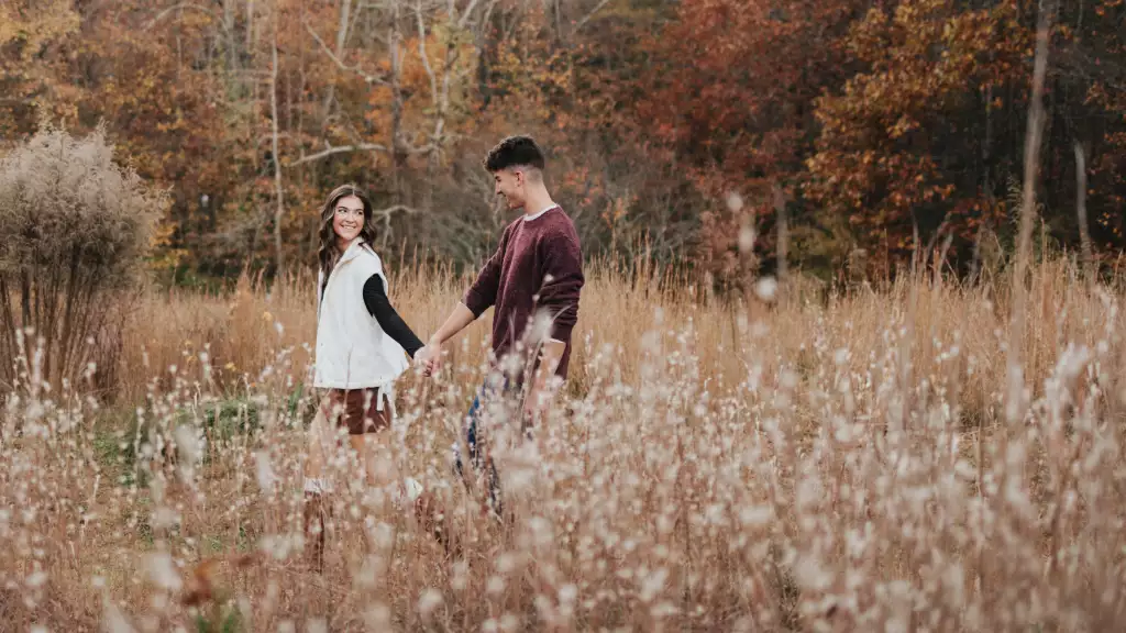 : Young couple holding hands and walking through a field of tall grasses and wildflowers with autumn trees in the background.