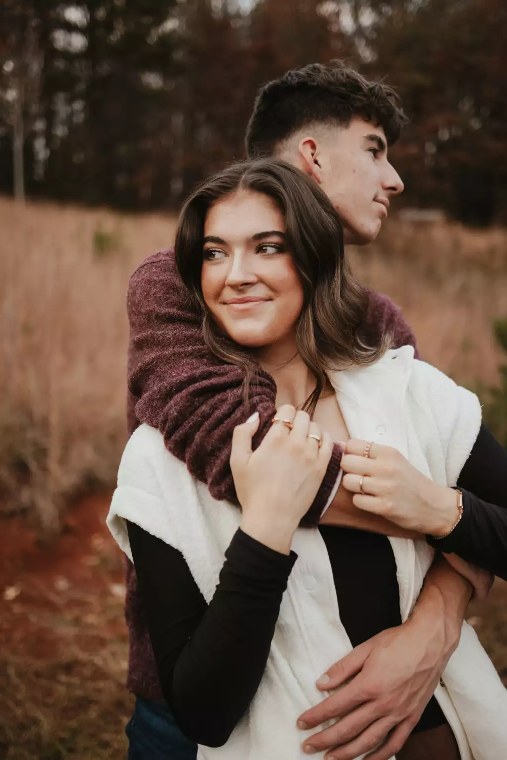 : Young couple embracing outdoors in autumn, woman smiling and looking to the side, man hugging her from behind and looking forward.