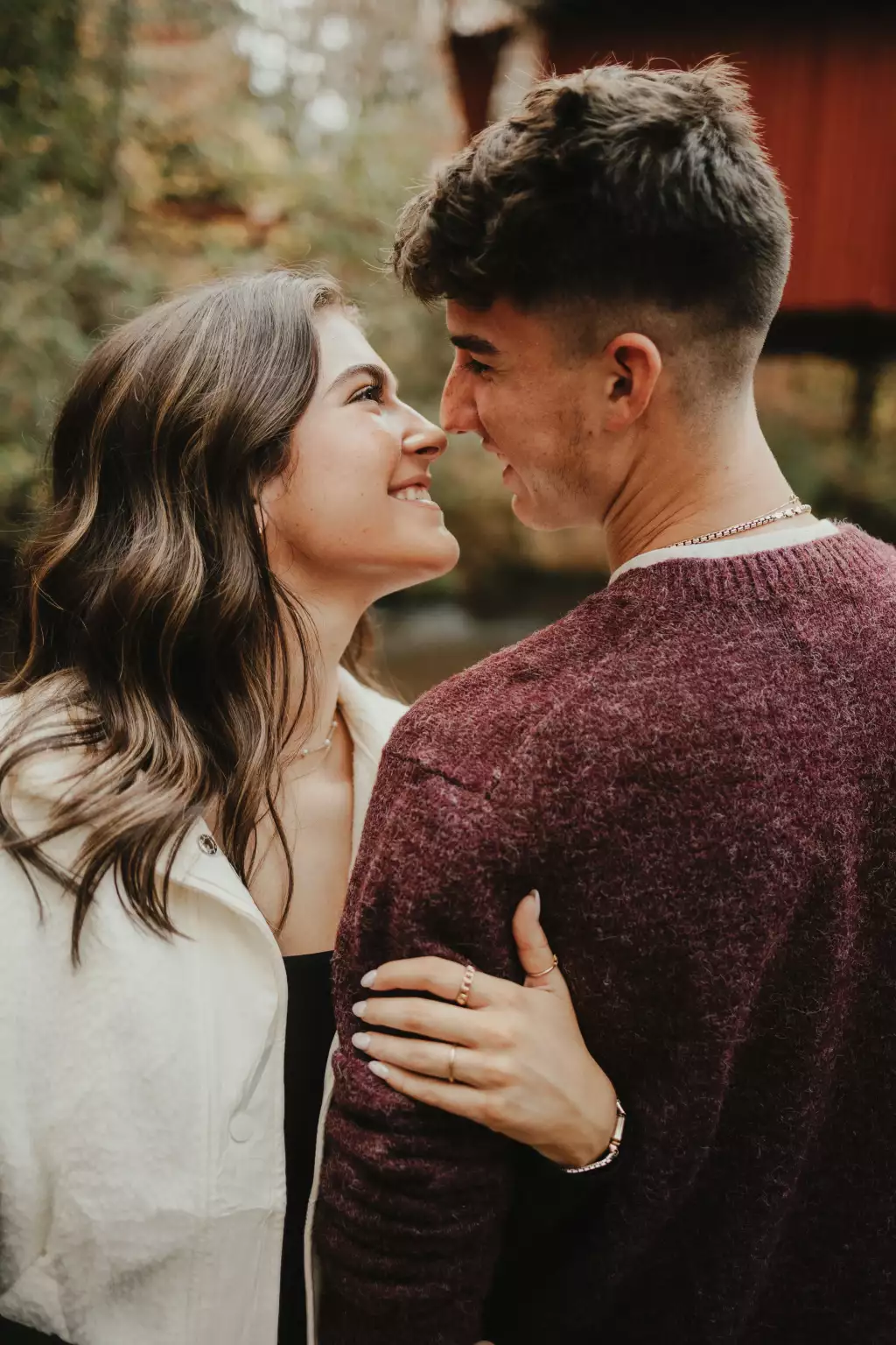 : Close-up of a young couple smiling and looking into each other's eyes outdoors, the woman wearing a white jacket and the man wearing a maroon sweater.